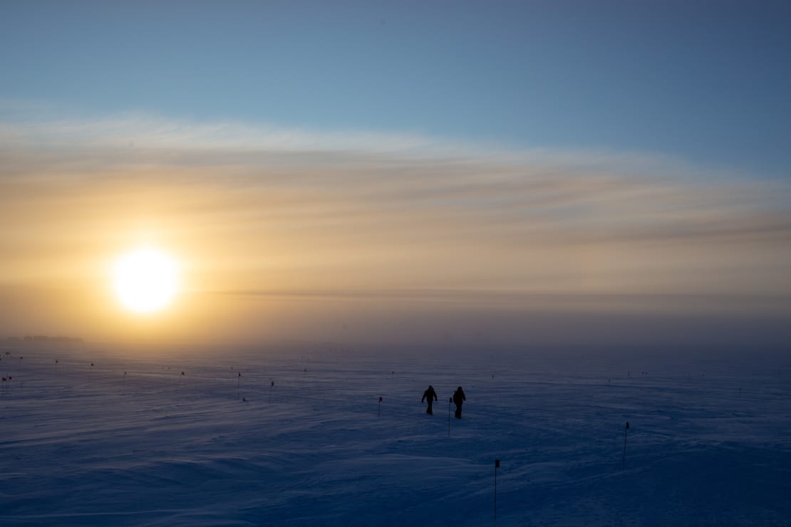 Large, bright sun low in the sky at the South Pole, with some wispy clouds and two people walking together in the distance.