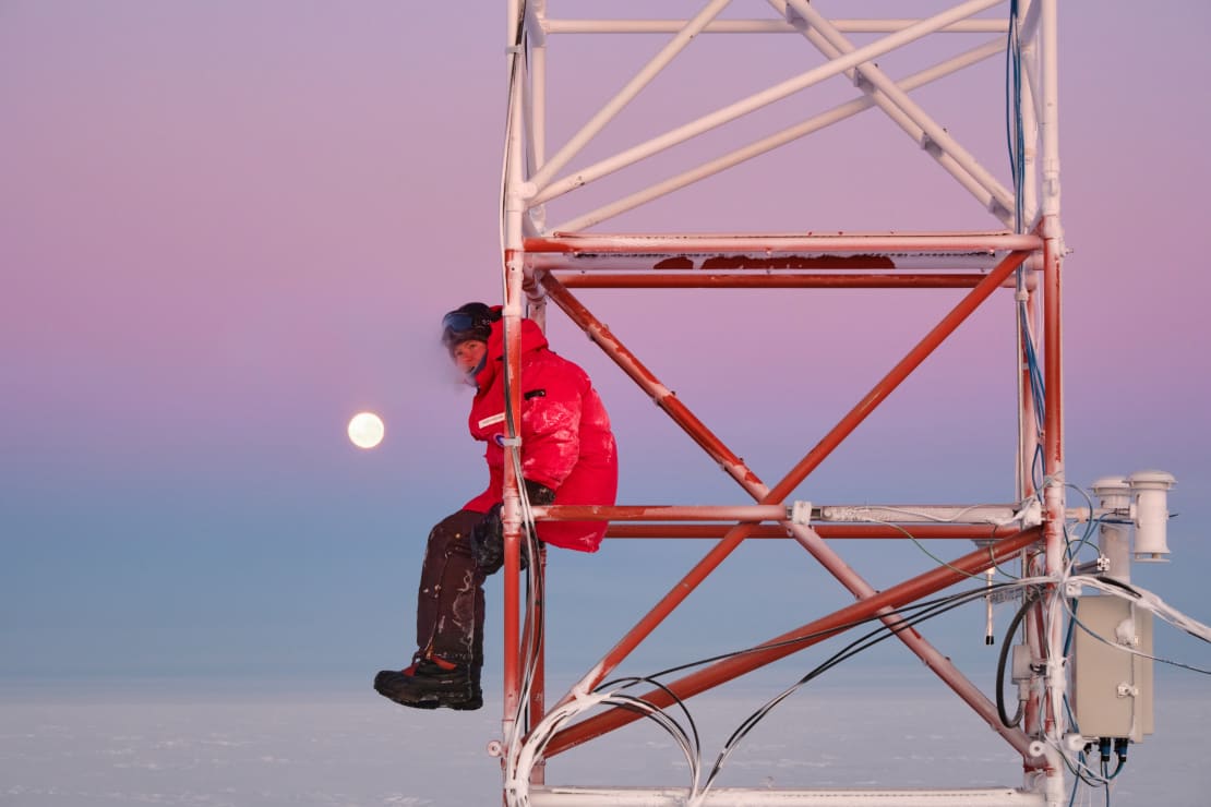 Person in red parka seen sitting on metal tower structure at the South Pole during sunset, beautiful pinks and purples in sky.