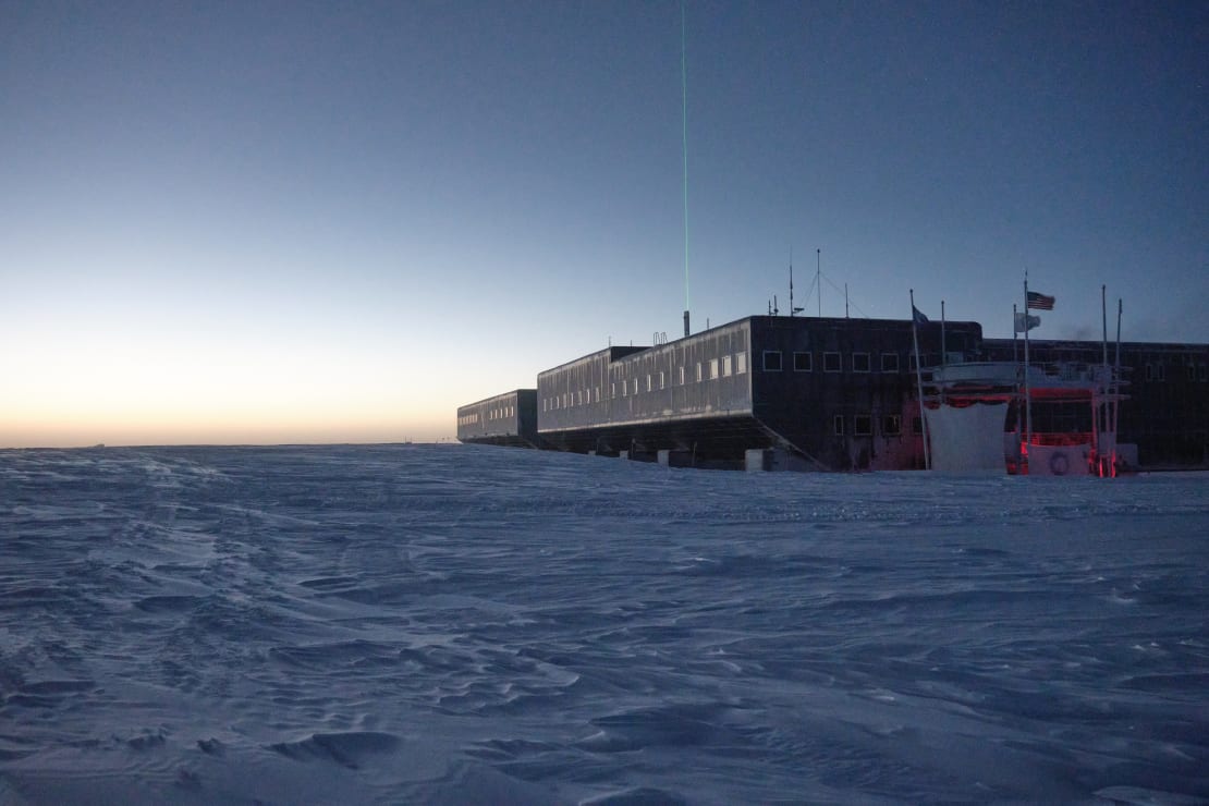 South Pole station post sunset, lingering light along horizon, and a LIDAR beam shooting up vertically into the sky from behind the station.