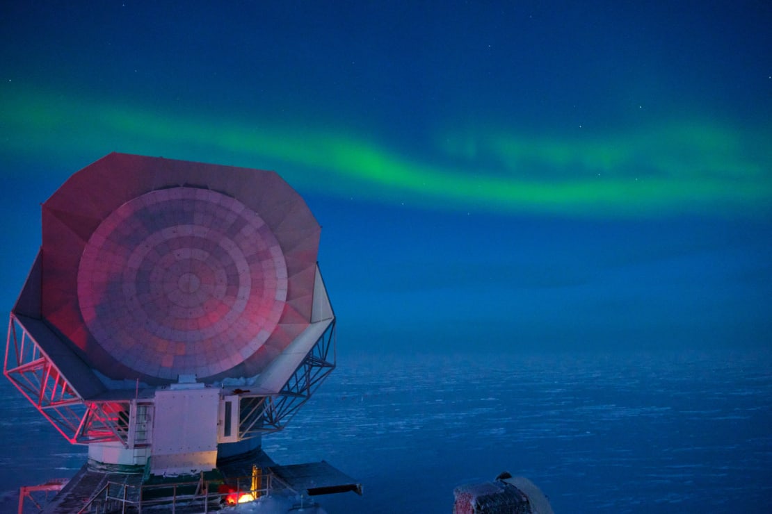 Bright green horizontal band of auroras across the sky with faintly lit South Pole Telescope in left foreground.