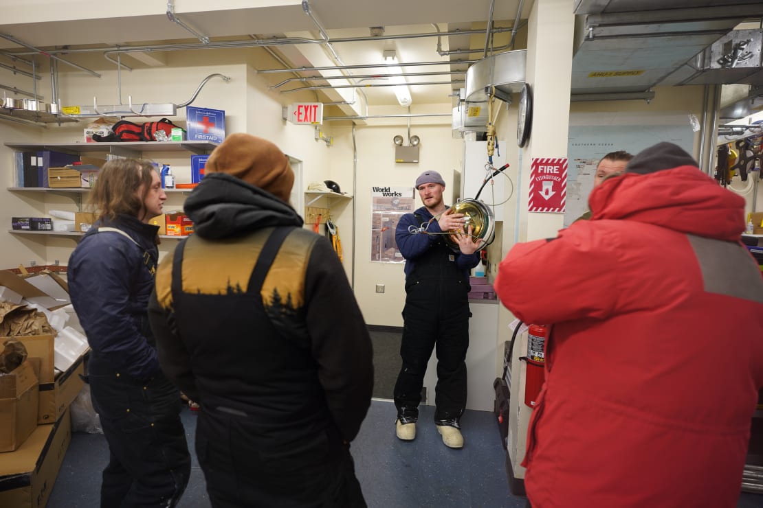 Small group gathered inside the IceCube Lab, around a winterover talking while holding an IceCube DOM.