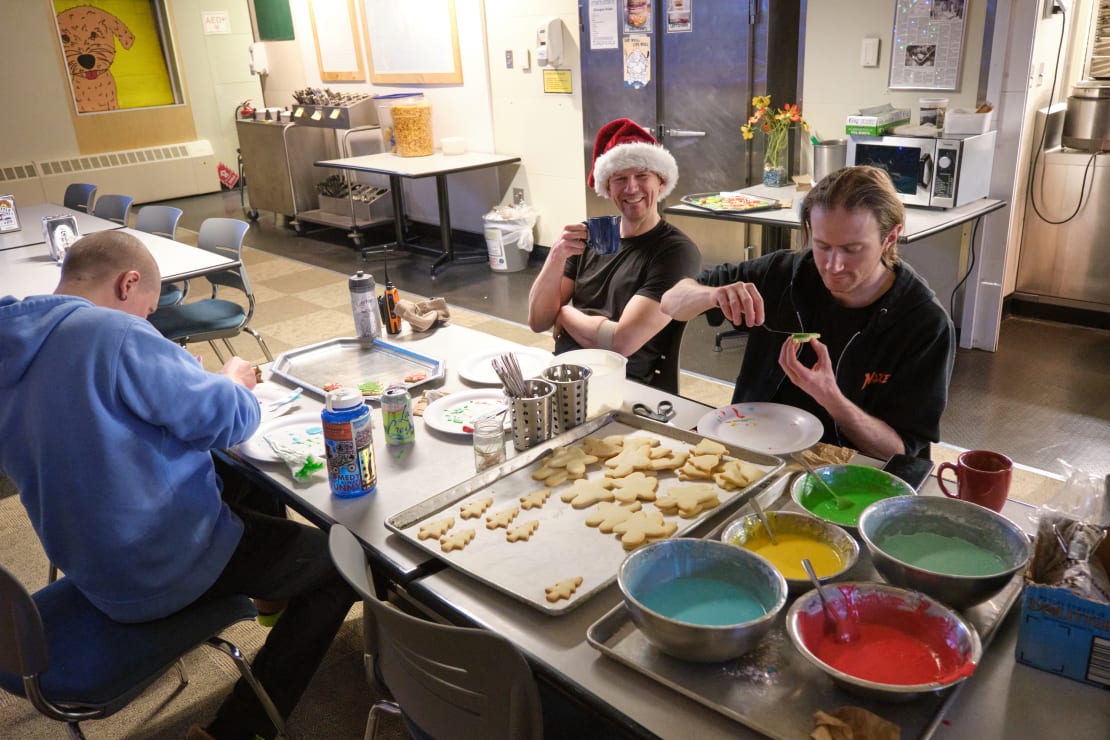 Three people decorating cut out cookies, seated at table, tray of undecorated cookies and large bowls of colored icing before them.