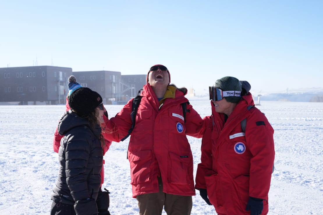 Close-up of three people out on the ice, station in the background, the middle person’s head thrown back in laughter.