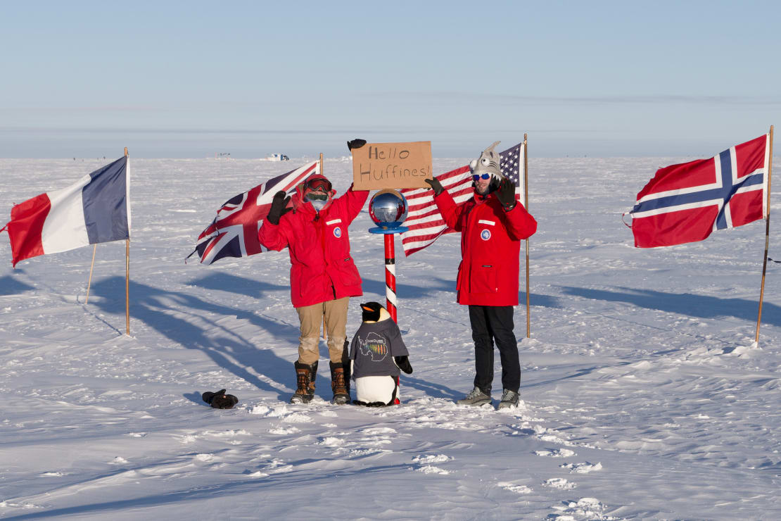 Two winterovers holding up greeting sign at the the ceremonial South Pole with a stuffed penguin doll at their feet.