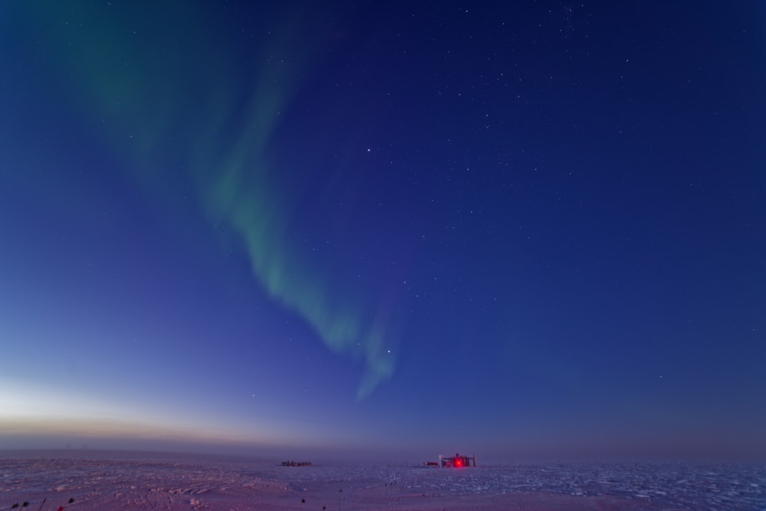 A narrow strip of light green aurora stretching up into the dark blue twilight sky at the South Pole, with the IceCube Lab appearing very small on the distant horizon.