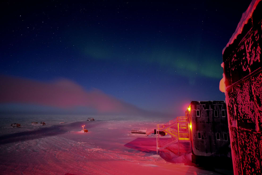 Winter view of auroras from behind the station, with the frosted edge of the building lit in red and off to the right.