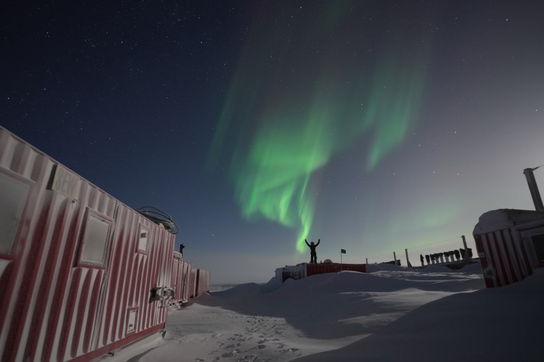 Person in shadow in the distance, with arms raised and standing on snowdrifted shipping container, under bright moon, a few wispy green auroras overhead.