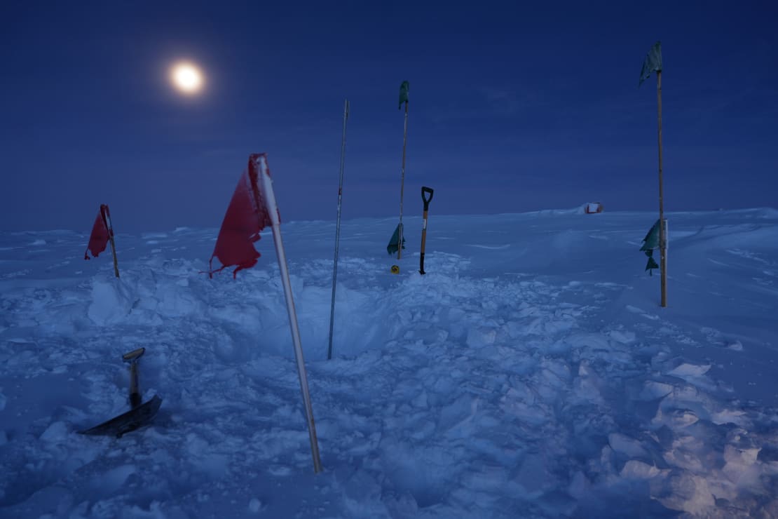 Shovel stuck upright into snow, flags around, moon overhead.