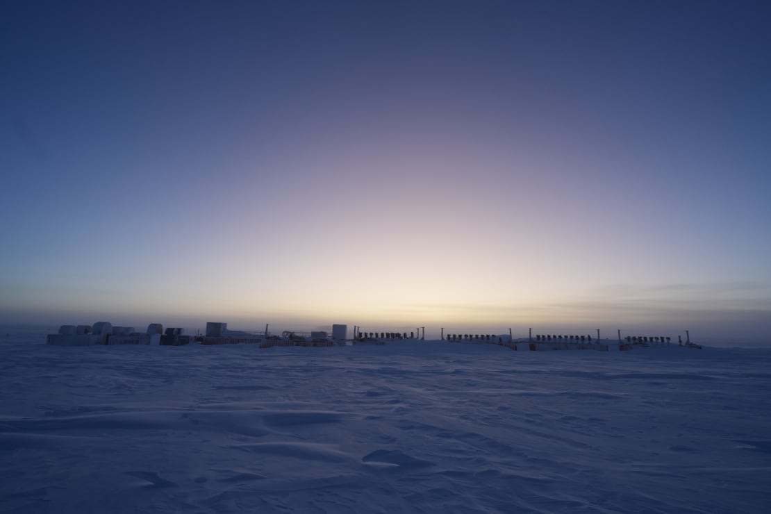 Light on horizon at South Pole twilight illuminating tops of lined up drill camp containers.