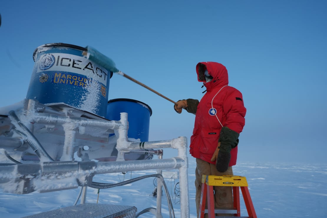 Winterover in red parka, standing on stepladder and brushing snow off scaffolded telescope apparatus.