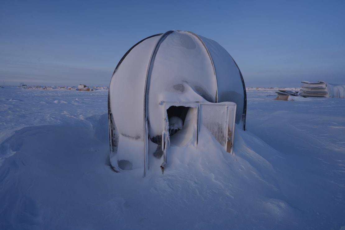 A round structure covered in frost, with open door and snow drifted in, at the South Pole storage berm.