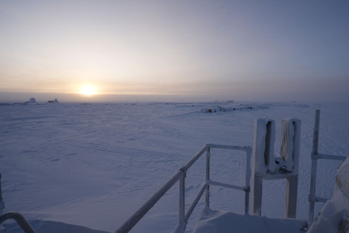 Sun just over the horizon at the South Pole, with the station and other buildings just visible in the distance.