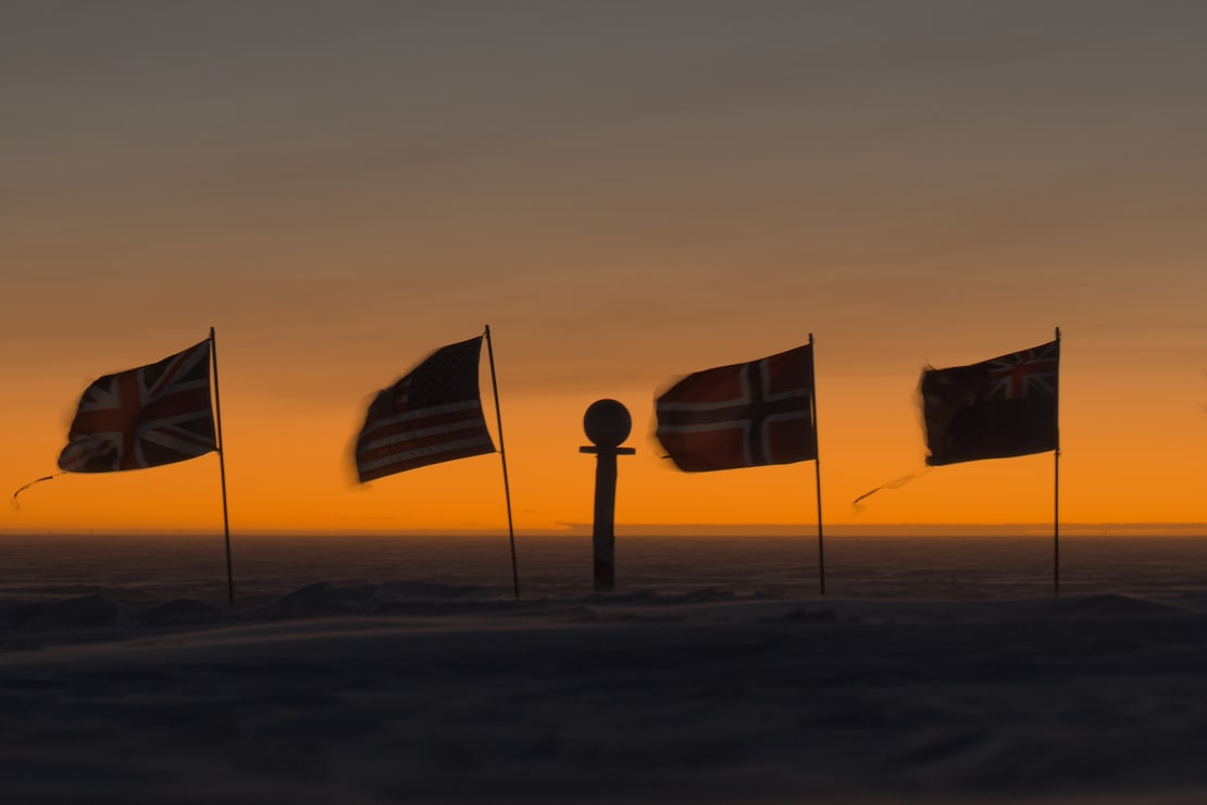 The marker and a few flags at the ceremonial Pole seen in shadow, with a bright orange sky along the horizon in background.
