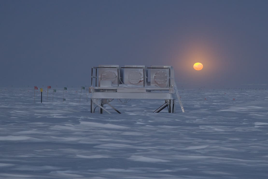 View of frosty outdoor structure at the South Pole, with moon behind it, low in sky over horizon, everything bathed in bluish hue.