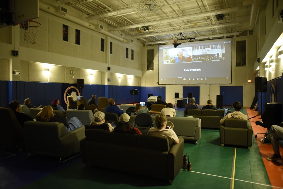 View from behind of theater-style seating arrangement in South Pole gym, with Zoom streaming on large screen.