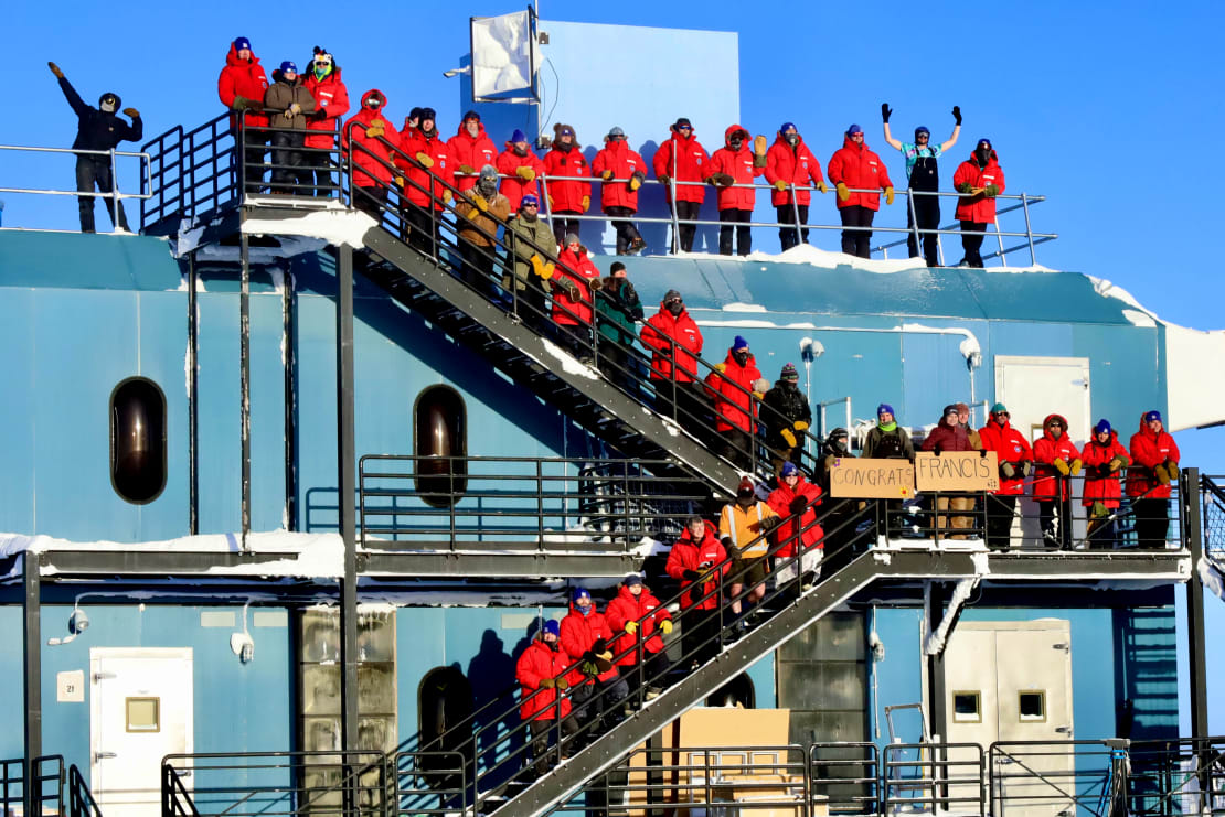 South Pole station winter crew in their red parkas all lined up for a group photo along the external staircase at the IceCube Lab.