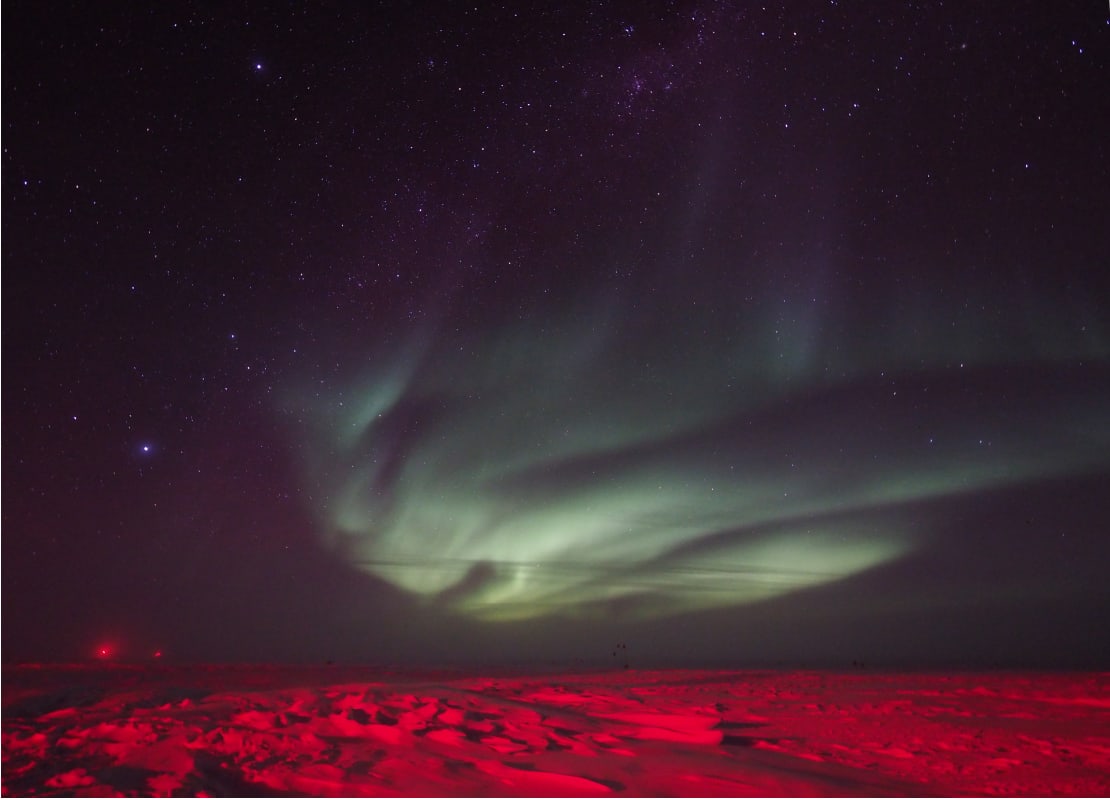 Swirling green auroras in dark sky, icy surface below lit up in red.