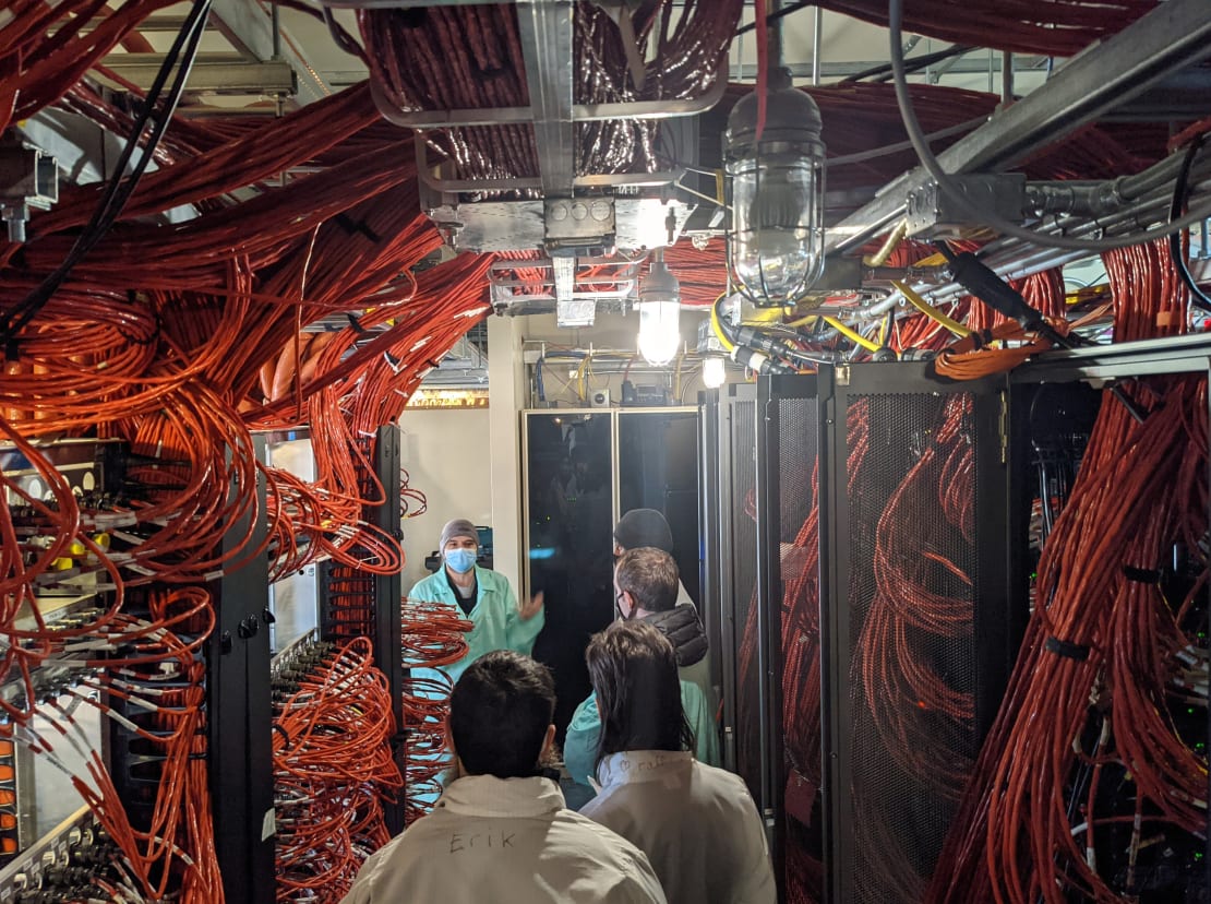 Masses of orange computer cables surrounding small group in lab coats listening to guide (wearing face mask) inside the IceCube Lab.