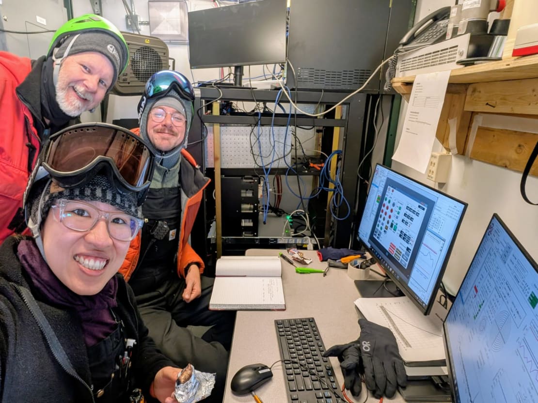 Three people in hard hats seated in front of computer monitors and smiling for the camera.