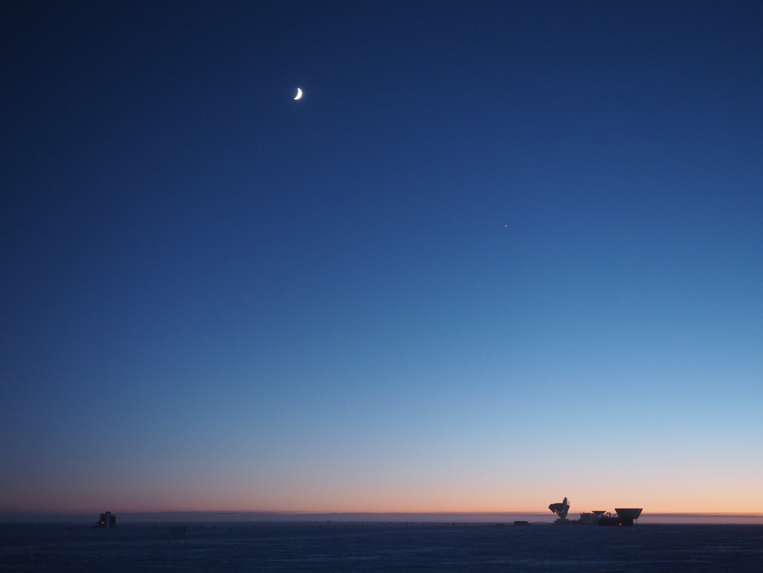 Twilight at the South Pole, with crescent moon high in sky and orange hues along horizon.