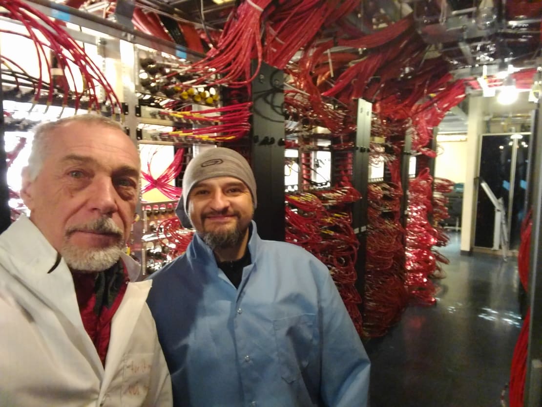 Two people close up in foreground, inside sever room of the IceCube Lab with lots of cables in background.