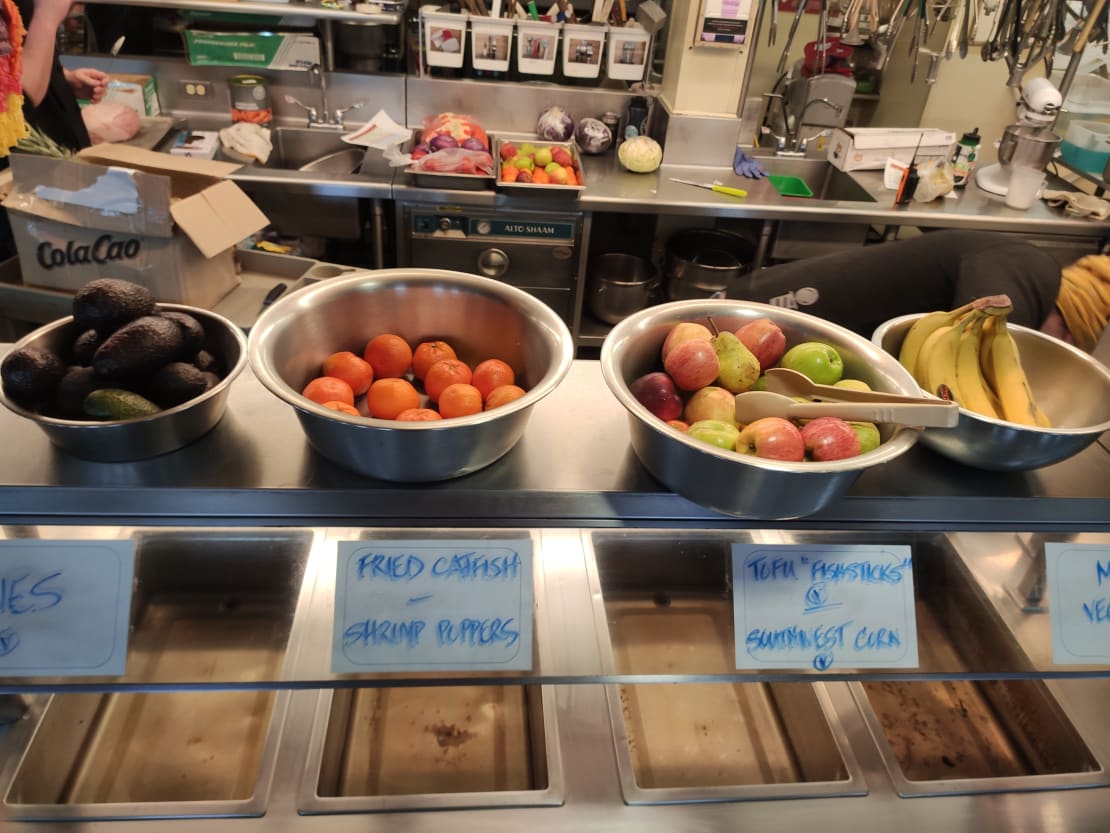 Bowls lined up on cafeteria shelf, each containing a single variety of fresh fruit.