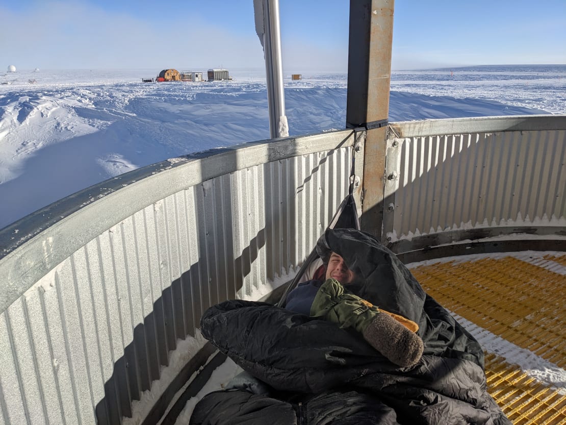 Smiling face and mittened glove all that show from a hammock stretched across outdoor platform at the South Pole station.