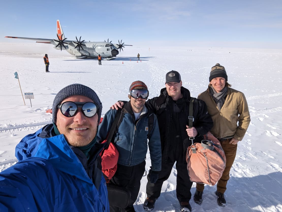 Selfie of group of four out on the ice with an LC-130 Hercules plane parked in the background.