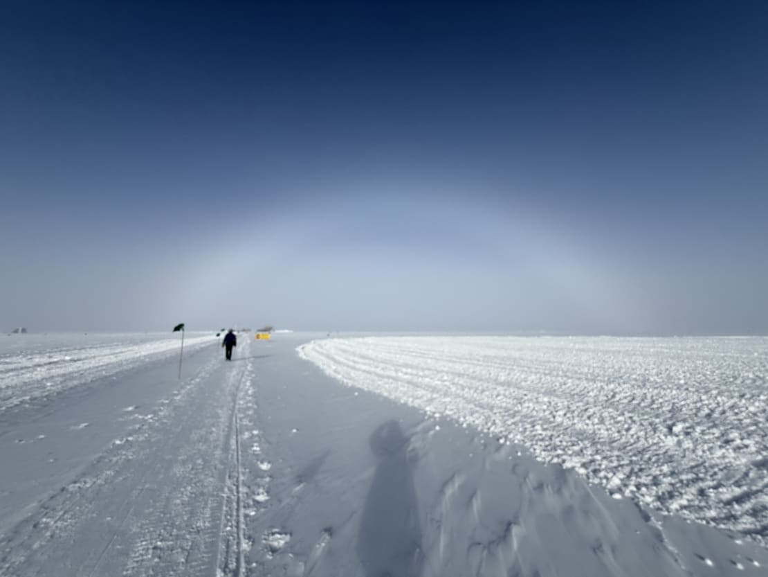 A white rainbow (or fogbow) on the horizon at the South Pole, with a person walking ahead in the distance.