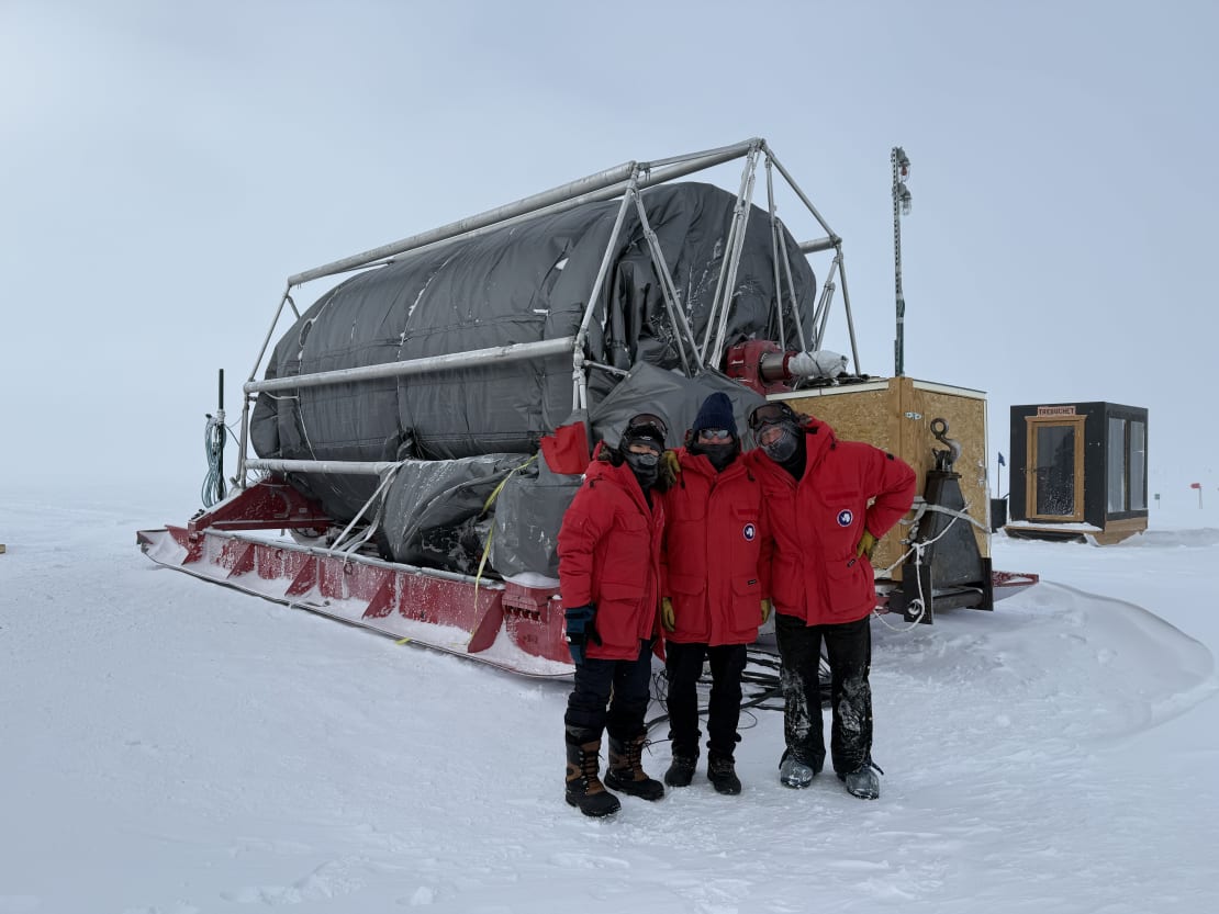 Three people in red parkas posing in front of large hose reel.