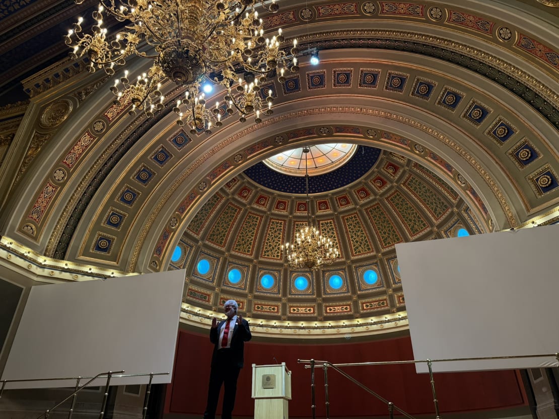 A person in a big room with chandeliers on stage with screens in the background.