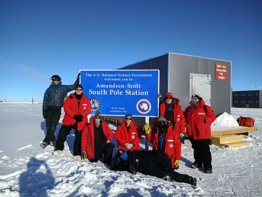 Group photo of eight people standing around the South Pole station sign.