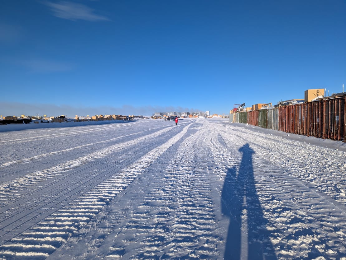 Long shadow of person on path as they photograph the South Pole station in the distance, storage containers off to the sides.