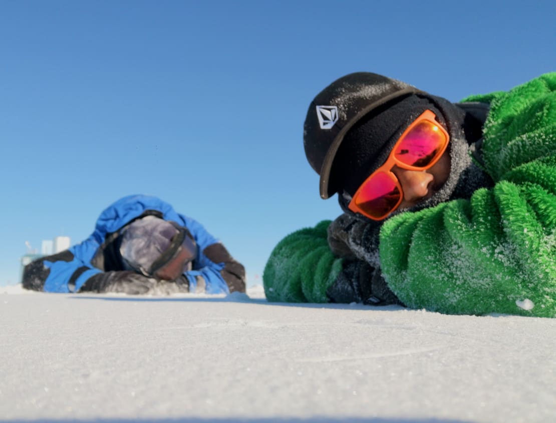 Low camera shot of two people lying down on the ice, one in sunglasses looking at the camera, the other with head down.