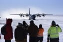 Group of people seen from behind in the foreground, large propellor plane approaching on ice in the background.