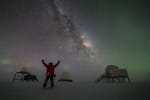 Person standing with arms up, and with three satellite dome structures in the background, and Milky Way, stars, and auroras in the sky.