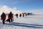 Group of people in red parkas walking out to an awaiting LC-130 plane on the South Pole skiway.