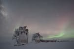 Wintry scene of two frosted and snow-covered IceCube drill towers with some faint auroras in the background.