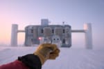 Focus on gloved hand holding a small paper penguin with blurry image of IceCube Lab in background.