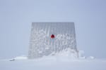 A huge reflector paneal at the South Pole, snowswept and with gray skies behind, and a winterover poking thru a small window about two-thirds the way up from the ground.