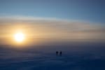 Large, bright sun low in the sky at the South Pole, with some wispy clouds and two people walking together in the distance.