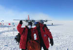 Two winterovers posing for the camera with an awaiting plane on the ice in the background under a blue sky.