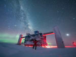 Winterover Josh with arms up, standing in front of IceCube Lab with auroras and Milky Way.