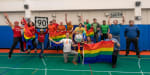 Group photo in the South Pole gym, with people in t-shirts the colors of the rainbow, and with arms up and holding rainbow flags.