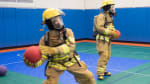 Two people in fire gear playing dodgeball in gym.]