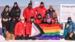 Group photo gathered around the geographic South Pole sign with pride flags.