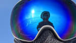 Close-up of winterover’s face covered by mask and googles, which are reflecting the parka-wearing photographer and the sun behind him.