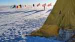 A Scott tent pitched on the ice at the South Pole, with the ceremonial Pole marker and some flags seen in the background.