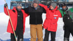 Group photo of four winterovers standing in front of the IceCube Lab under blue sky.