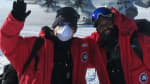 Two winterovers posing for the camera with an awaiting plane on the ice in the background under a blue sky.
